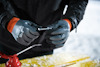 Person with black gloves holding a small metal tool, standing at a yellow wooden surface.
