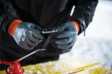 Person with black gloves holding a small metal tool, standing at a yellow wooden surface.