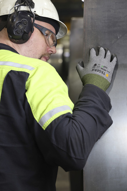 Person med hjälm och hörselskydd inspekterar metallplåt med hand iklädd en handske från Tegera.