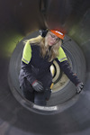 En person med skyddshjälm och hörselskydd inspekterar insidan av ett stort rör eller tunnel, iförd arbetskläder.