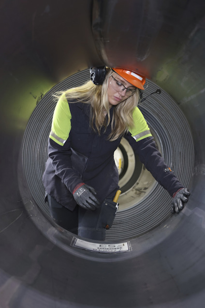 En person med skyddshjälm och hörselskydd inspekterar insidan av ett stort rör eller tunnel, iförd arbetskläder.