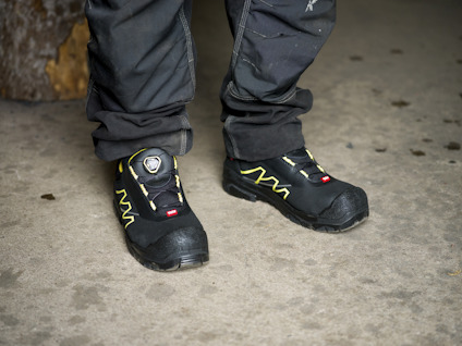 Person med svarta arbetsbyxor och svarta skyddsskor med gula detaljer står på betonggolv.