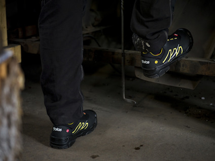 Svart- och guldfärgade skyddsskor med logotyp, används av person i svart arbetsklädsel.