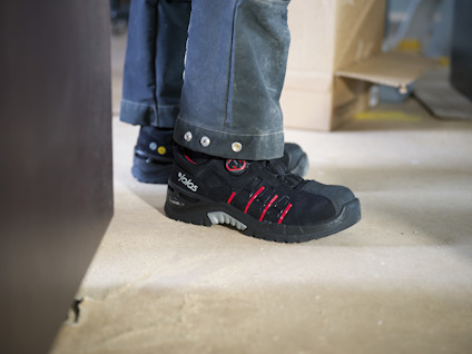 Arbetsskor med röd detalj och byxor, kartong i bakgrunden på betonggolv.