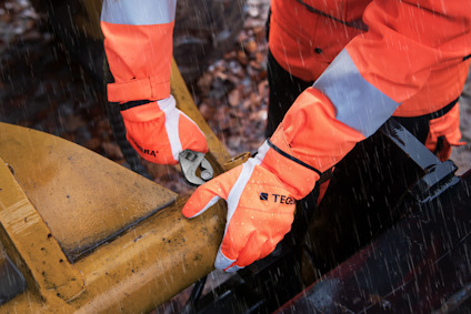 Person i orange arbetskläder med handskar använder verktyg på gul maskindel i regn.