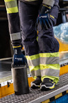 Person i arbetskläder med reflexer, skyddshandskar och skor, håller ett mörkt metallföremål och står på en plattform.