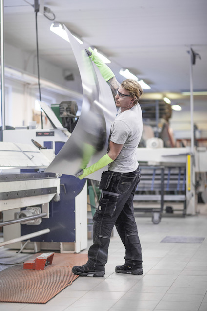 Person i industriell miljö lyfter ett stort metallark iklädd skyddshandskar. Flera maskiner syns i bakgrunden.
