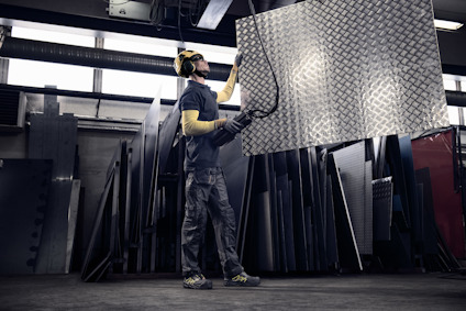 Person som hanterar stort metallstycke i fabriksmiljö, omgiven av fler metallplattor och industriell utrustning.