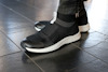 Svarta sneakers med vit sula på svart kakelgolv. En person bär skorna.