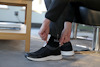 Person som drar upp sina byxor för att visa svarta strumpor och svarta sneakers med vita sulor, sittande inomhus.