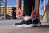 Person böjer sig för att knyta svarta sneakers utomhus, en annan person promenerar i bakgrunden.