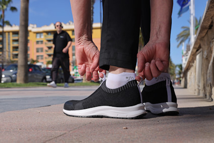 Person böjer sig för att knyta svarta sneakers utomhus, en annan person promenerar i bakgrunden.