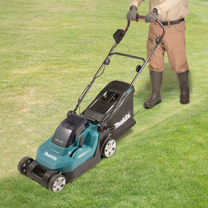 Gräsklippare av märket Makita på gräsmatta, person i stövlar och beige kläder skjuter den.