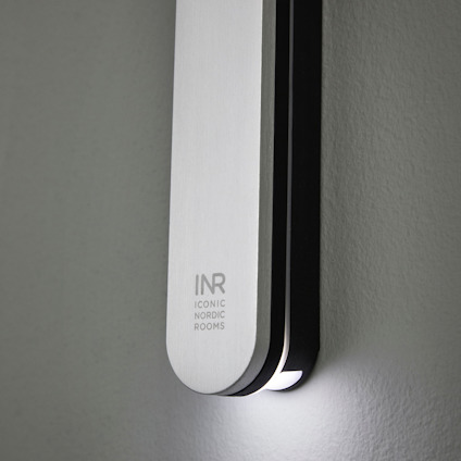 Väggmonterad armatur i metall med texten "INR Iconic Nordic Rooms", avger ljus nedåt.