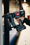 En person håller i en svart och grå spikpistol med märket Senco nära en vägg.