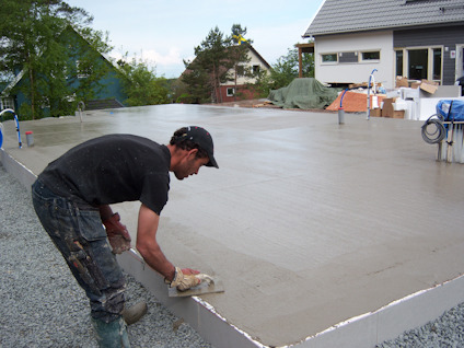 En person jämnar till ytan på en nyligen gjuten betongplatta framför ett hus under byggnation.
