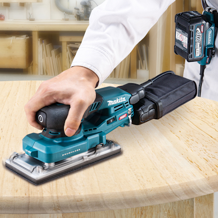 En person slipar trä med en blå och svart Makita sladdlös slippapper-maskin med ett batteripack.