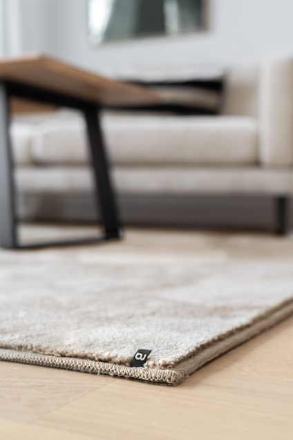 Beige matta på ljust trägolv, soffbord och soffa i bakgrunden. Avslappnad interiörstil.