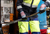Person i skyddsoverall och röda handskar håller ett metallverktyg på byggarbetsplats.