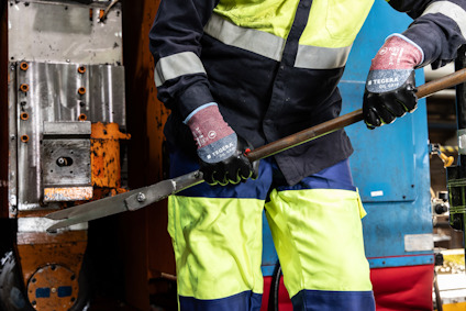 Person i skyddsoverall och röda handskar håller ett metallverktyg på byggarbetsplats.