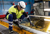 Person med skyddshjälm och skyddsglasögon hanterar en stor skiva glas i industriell miljö.