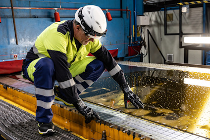 Person med skyddshjälm och skyddsglasögon hanterar en stor skiva glas i industriell miljö.