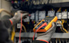 Person med skyddshandskar använder en orange multimeter för att mäta spänning i elektrisk panel.