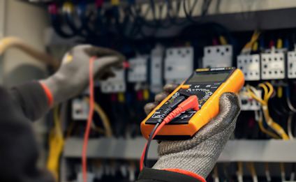 Person med skyddshandskar använder en orange multimeter för att mäta spänning i elektrisk panel.
