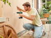 Man in a yellow shirt and jeans using a cordless drill on an outdoor wall next to a garden chair and plant.