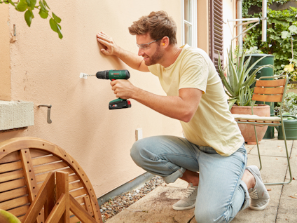 Man in a yellow shirt and jeans using a cordless drill on an outdoor wall next to a garden chair and plant.