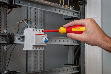 Hand som använder en röd-gul skruvmejsel på en vit elektrisk komponent i ett metallställ.