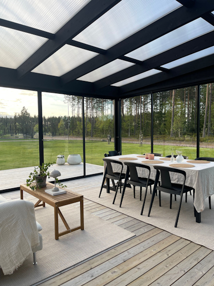 Inglasat uterum med svart ram, trägolv, och möbler med vit duk. Skog och gräsmatta utanför.