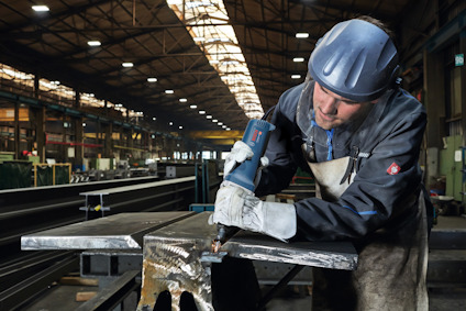 Person i hjälm och skyddskläder slipar metall i en stor industrihall.