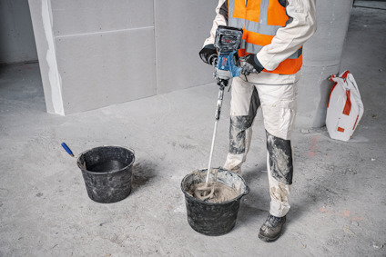 Arbetare i vit och orange skyddsutrustning blandar material i en hink med en mixer, betonggolv och väggskivor i bakgrunden.