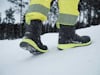 Person i gula byxor och svarta vinterkängor går i snöigt landskap med träd i bakgrunden.