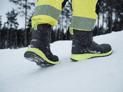 Person i gula byxor och svarta vinterkängor går i snöigt landskap med träd i bakgrunden.
