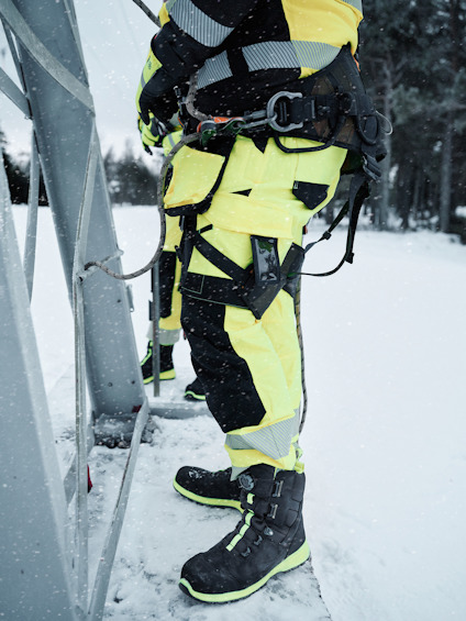 Person klädd i gul arbetsklädsel och svarta vinterkängor som arbetar utomhus vid en metallstruktur i snöigt väder.