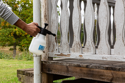 En person sprayar en träveranda med en vit sprayflaska. Verandan har avskavd vit färg.