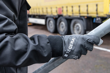 En person i arbetskläder håller i ett långt metallrör med en handske märkt "TEGERA". I bakgrunden syns en gul trailer.