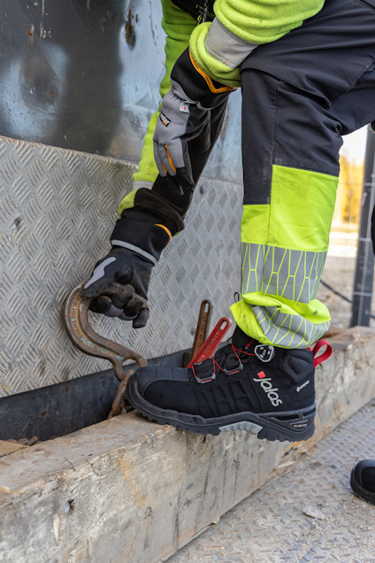 Person i arbetskläder och handskar böjer sig ner vid en metallplatta, bär skyddskängor och reflexförsedd overall.