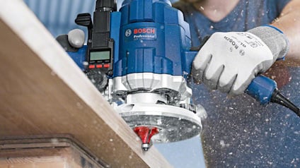 En hand håller i en blå Bosch Professional fräs, som bearbetar en kant av trä. Brus och träspån syns i luften.