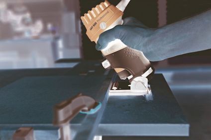 Hand håller en blå och vit handöverfräs mot en träskiva på ett bord med ljusa trämöbler i bakgrunden.