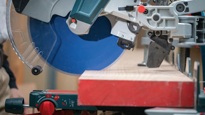 Kap- och gersåg med blått sågblad skär genom träplanka, omgiven av verkstadsutrustning.