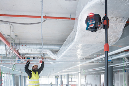 En person i skyddsväst installerar metallrör i ett industriellt innertak med ventilationskanaler och en laserpassare.