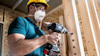 Person med skyddshjälm och mask borrar hål i träbalkar inomhus.