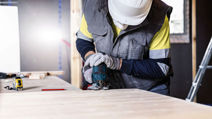 Arbetare i skyddshjälm och reflexväst borrar i trä, verktyg och stege i bakgrunden.