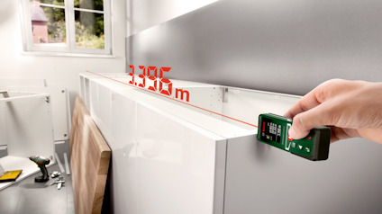 Hand använder digitalt lasermätverktyg. Visad mätning: 3,336 m. Byggmaterial och verktyg synliga i bakgrunden.