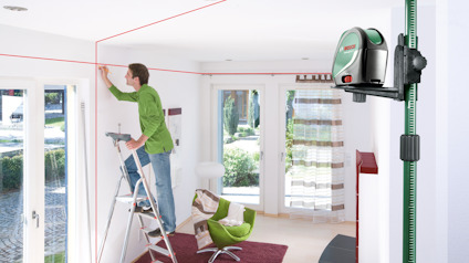 En person använder en stege och lasernivå för att anpassa design och renovering i ljust rum med stora fönster.