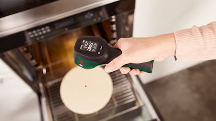 Hand håller en termometer som visar 177,3°C ovanför en ugn med en pizzasten inuti.