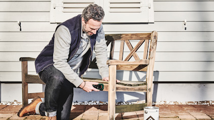 Man knäböjer vid en träbänk utomhus, håller en liten enhet nära bänken. Färgburk står på marken.